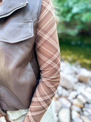 A close-up outdoor shot of a person wearing a brown faux-leather vest over a sheer long-sleeve plaid shirt in tan and brown tones, with a blurred rocky stream and greenery in the background.