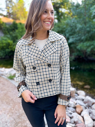 A woman wearing a cream and black gingham double-breasted blazer with brown trim, standing outdoors near a stone path and water.