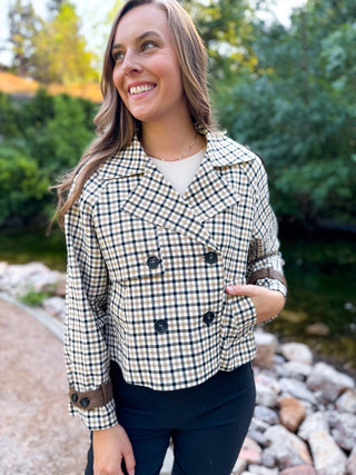 A woman wearing a cropped, double-breasted plaid jacket with brown elbow patches and button details, standing outdoors near a stone path and water.