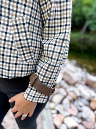 A close-up of a person wearing a brown and beige plaid flannel shirt with a brown rolled cuff, paired with black pants, set against a blurred outdoor background of rocks and greenery.