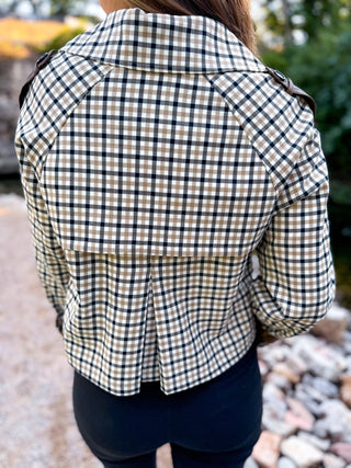 A person viewed from behind wearing a cropped plaid button-up shirt with dark shoulder epaulets, paired with dark pants, set against an outdoor background of rocks and greenery.