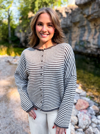 A woman wearing a gray and white horizontally striped knit cardigan with a button-down front, standing outdoors in front of a rocky, wooded background.