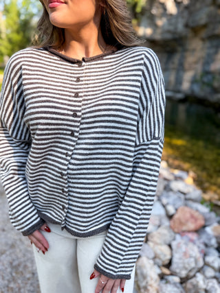 A woman wearing a brown and gray striped button-up cardigan sweater, photographed outdoors near a stone wall and water.
