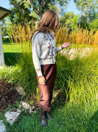 A woman stands outdoors in a garden wearing a cream knit cardigan with brown and black embroidered patterns, paired with brown pants and dark boots.