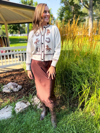 A woman stands outdoors in a grassy area next to a covered patio, wearing a cream knit cardigan with brown and red western-style embroidery, a rust-brown midi skirt, and dark brown boots.