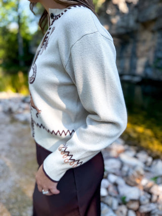 A person wearing a cream knit sweater with brown embroidered geometric and animal motifs, photographed outdoors near rocks and water.