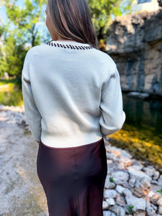 A woman viewed from behind wearing a light gray knit sweater with a dark brown decorative collar and a dark burgundy skirt, standing outdoors near a stone path and water.