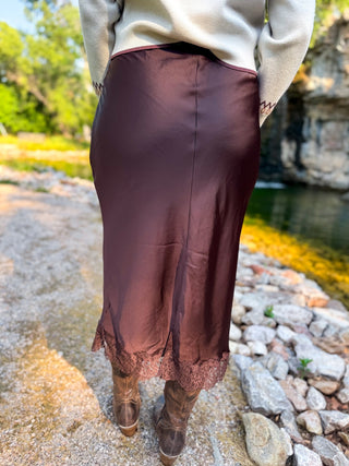 A person standing outdoors by a rocky stream wearing a long satin-like brown skirt with lace trim, a cream sweater, and brown boots.
