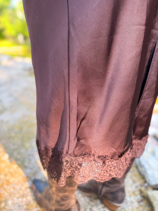 Close-up of the lower portion of a dark brown satin or silk skirt with a lace hem, worn with brown boots outdoors.