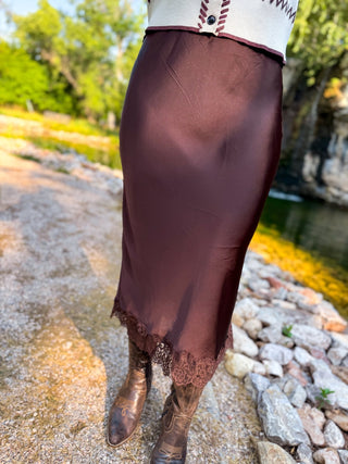 A person outdoors wearing a dark brown satin midi skirt with a lace hem, paired with brown cowboy boots and a cream sweater.