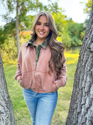 A smiling woman stands outdoors between two tree trunks, wearing a dusty pink zip-front jacket over a green plaid button-up shirt and light blue jeans.