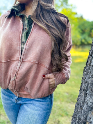 A woman wearing a dusty rose zip-up fleece jacket over a green plaid shirt and blue jeans, standing outdoors next to a tree trunk.