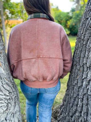 A person viewed from behind wearing a dusty rose pink cropped sweatshirt with a green plaid collar, standing outdoors between two large tree trunks.