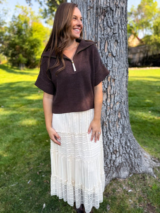A woman stands outdoors next to a large tree trunk on a grassy lawn, wearing a dark brown short-sleeve ribbed knit top with a half-zip collar and a long cream pleated skirt.