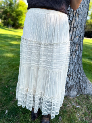 An off-white tiered maxi skirt with textured horizontal bands and ruffled edges, worn by a model standing outdoors next to a tree.