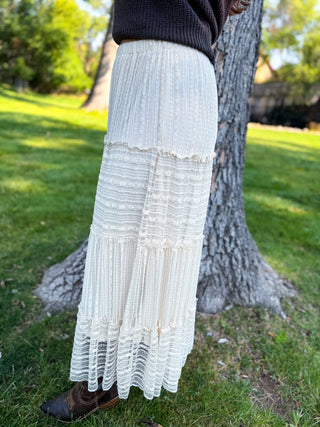 A person outdoors wearing a long cream pleated maxi skirt with a dark ribbed top and brown boots, standing beside a tree on a grassy lawn.