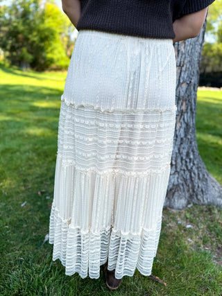 An off-white tiered maxi skirt with textured horizontal bands and ruffled hems, worn by a model standing outdoors next to a tree on a grassy lawn.