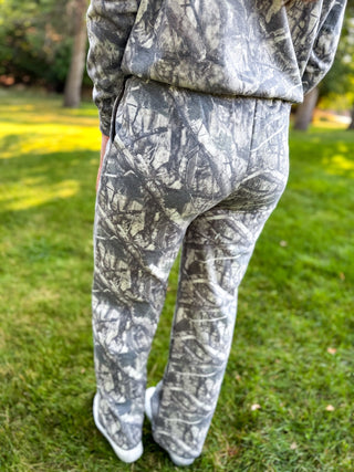 A person standing outdoors on grass, viewed from the back, wearing a matching camouflage tracksuit with a gray, black, and off-white woodland print.