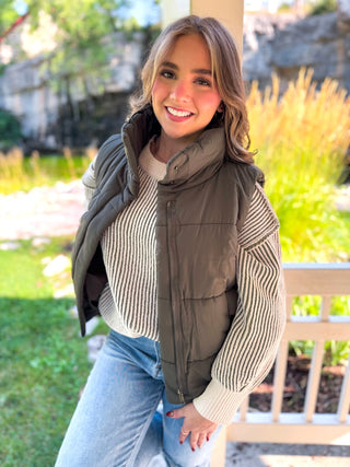A woman wearing an olive green puffer vest over a cream and black striped knit sweater, paired with light blue jeans, standing outdoors in front of a porch post and garden background.