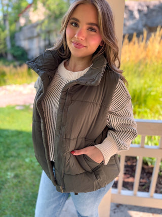 A woman wearing an olive green puffer vest over a cream ribbed knit sweater and light blue jeans, standing outdoors in front of a white wooden fence and greenery.