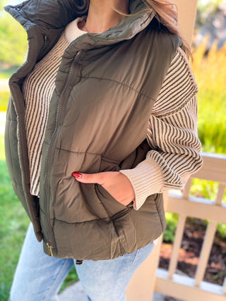 A woman wearing an olive green puffer vest over a beige and brown striped knit sweater, paired with light blue jeans, standing outdoors near a wooden bench.
