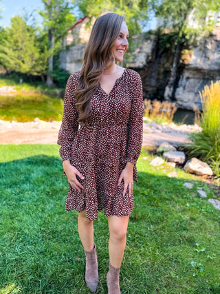 A woman wearing a brown and white patterned long-sleeve mini dress and taupe ankle boots, standing outdoors on a green lawn with trees and a stone wall in the background.