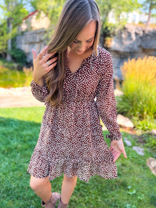 A woman wearing a brown and white speckled long-sleeve dress, standing outdoors on a grassy lawn with garden plants and a stone wall in the background.