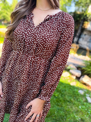 A woman wearing a brown long-sleeve dress with a white ditsy dot pattern, photographed outdoors in a sunny garden setting.