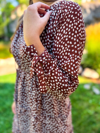 A close-up of a person wearing a brown long-sleeve dress with a white speckled or polka-dot pattern, standing outdoors on a grassy lawn with a blurred background.