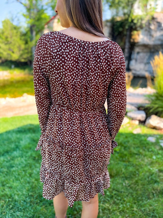 A woman wearing a long-sleeve brown dress with a white speckled pattern, shown from the back in an outdoor garden setting.