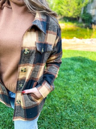 A person outdoors wearing a brown, beige, and navy plaid button-front overshirt over a ribbed brown sweater, with grass and trees in the background.