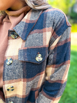 Close-up of a person wearing a plaid button-front overshirt over a pink knit top, with a blurred outdoor green background.