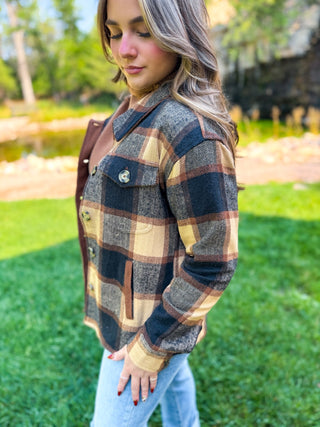 A woman wearing a brown, beige, and black plaid flannel shirt jacket, standing outdoors on a green lawn with trees in the background.