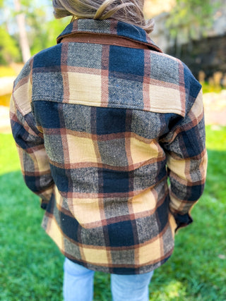 A person viewed from behind wearing a long-sleeve plaid flannel-style shirt jacket in navy, tan, and brown, standing outdoors on grass.