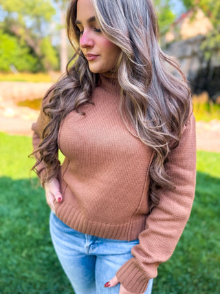 A woman wearing a ribbed knit mock neck sweater in a warm camel brown color, paired with light blue jeans, standing outdoors on a grassy lawn.