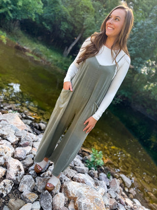 A woman wearing an olive green sleeveless jumpsuit over a white long-sleeve shirt, standing on rocks next to a river with trees in the background.