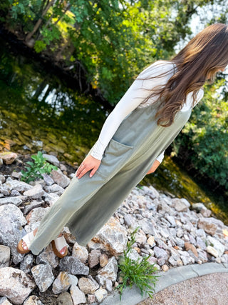 A woman wearing a sage green corduroy pinafore dress over a white long-sleeve top, standing on a rocky path next to a stream with lush green trees in the background.