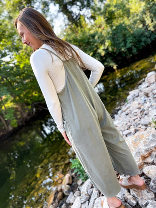 A woman standing on rocks by a river wearing an olive green wide-leg overall jumpsuit over a cream long-sleeve top.