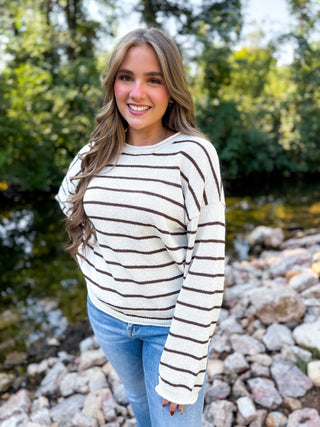 A smiling woman standing outdoors near a rocky stream, wearing a cream knit sweater with thin brown horizontal stripes and light blue jeans.