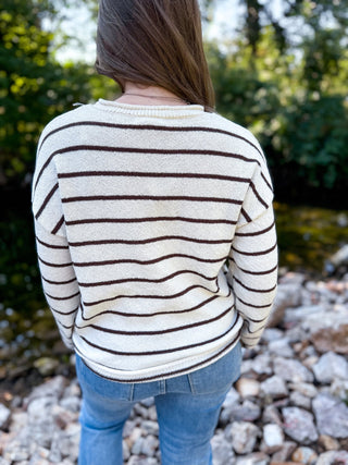 A person viewed from behind wearing a cream knit sweater with dark brown horizontal stripes and blue jeans, standing outdoors near rocks and greenery.