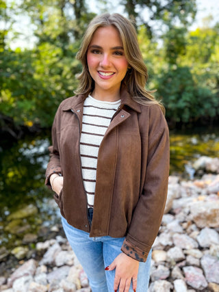A smiling young woman stands outdoors near a rocky stream with green trees in the background, wearing a brown suede-like jacket over a white and brown striped shirt with light blue jeans.