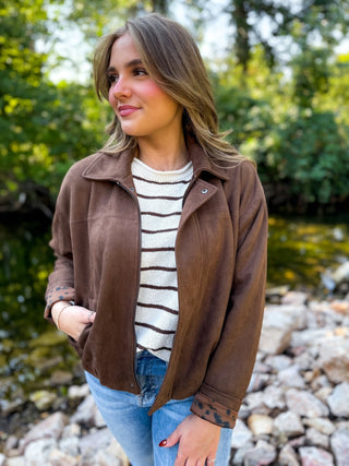 A woman wearing a brown suede jacket over a cream and brown striped sweater, blue jeans, and a bracelet, standing outdoors on a rocky area with green trees and water in the background.