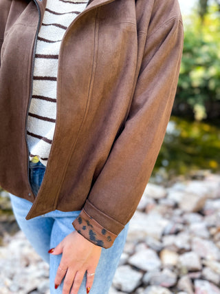 A person wearing a brown suede-like zip-front jacket over a cream and brown striped sweater, paired with light blue jeans, standing outdoors near rocks and water.