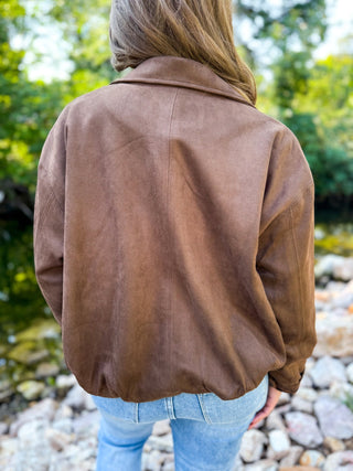 A person viewed from behind wearing a brown suede or faux-suede jacket outdoors near rocks and greenery, paired with light blue jeans.