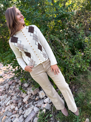 A woman wearing a cream knit cardigan with brown and beige argyle diamonds and gold buttons, paired with beige pants, standing outdoors on a rocky grassy area with green foliage in the background.