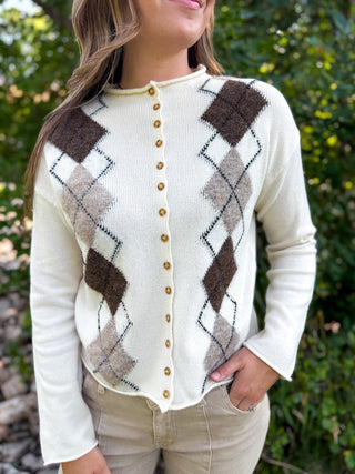 A woman wearing a cream-colored knit cardigan sweater with brown and beige argyle diamond patterns and a row of gold buttons, paired with beige pants, set against a blurred green outdoor background.