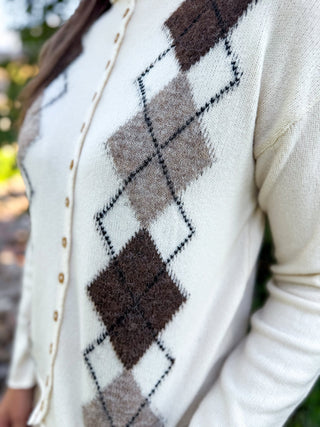 Close-up of a person wearing a cream knit cardigan with an argyle pattern in dark brown, taupe, and black, featuring gold-tone buttons down the front.