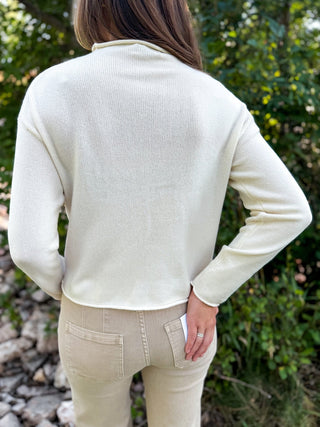 A woman viewed from behind wearing a cream ribbed long-sleeve mock neck top and beige pants, standing outdoors in front of a blurred green foliage background.