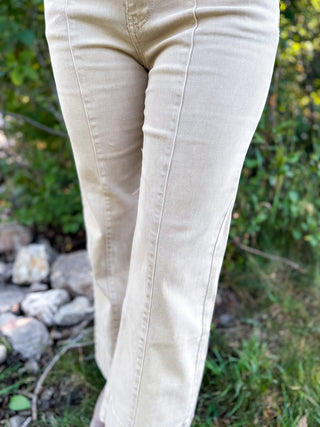 Close-up of a person wearing light beige straight-leg or bootcut jeans outdoors on grass with rocks and greenery in the background.