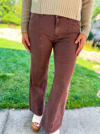 A person wearing brown wide-leg cropped pants with a ribbed beige sweater, standing outdoors on a concrete path with green grass in the background.